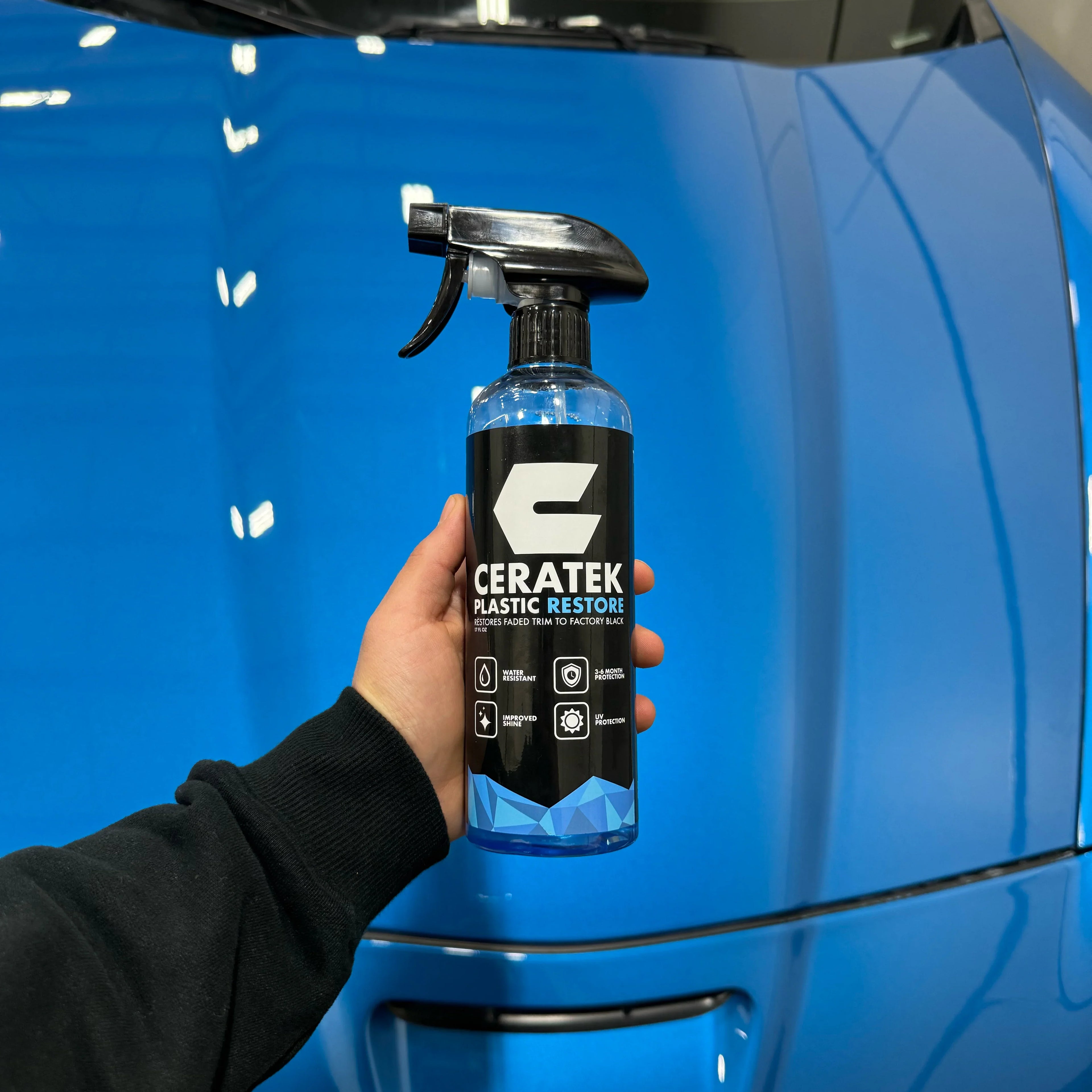 Hand holding Ceratek Plastic Restore spray in front of a shiny blue car hood.
